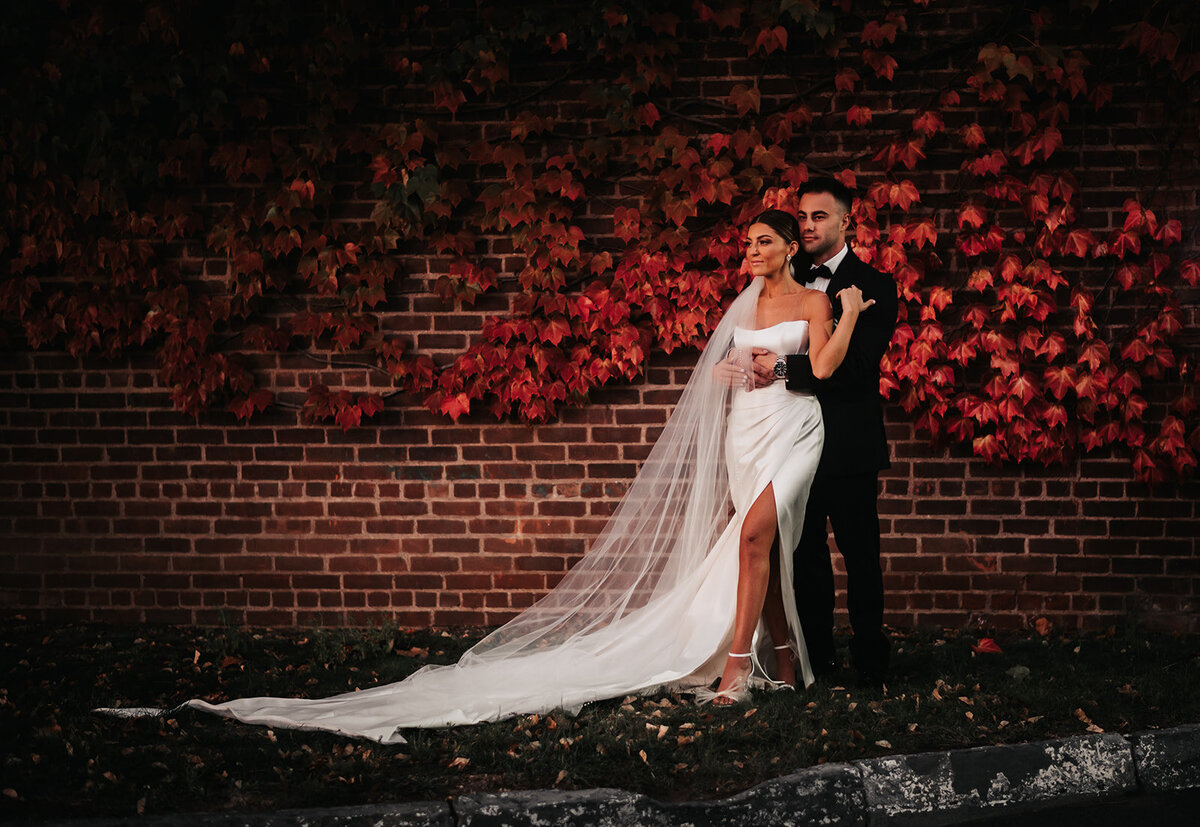 bride and groom share an embrace in wedding attire with red leaves on wall behind them at their new haven lawn club wedding photo by cait fletcher photography