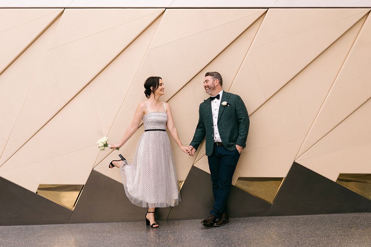 Brisbane Registry Office wedding couple holding hands and walking playfully in front of a geometric wall, with the bride lifting her heel and holding a bouquet.