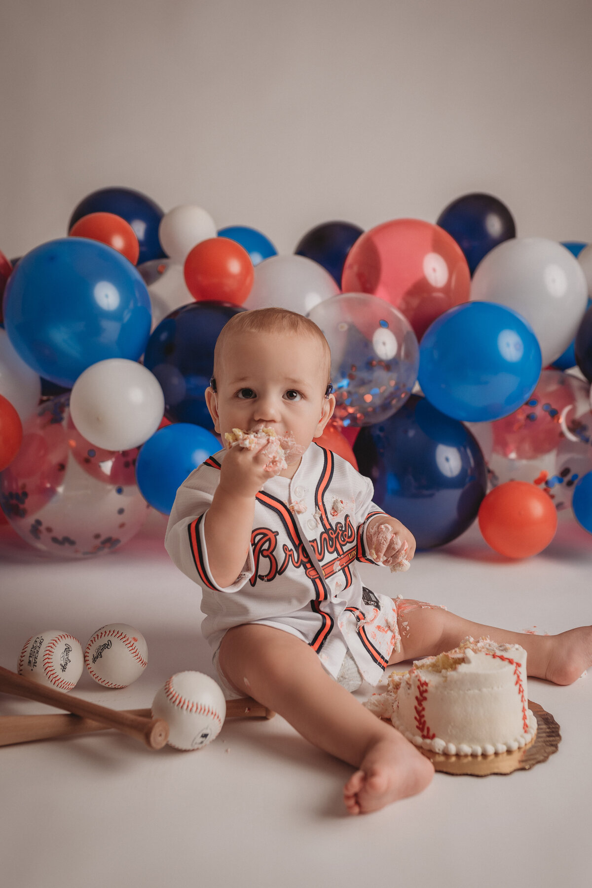 Atlanta Braves themed cake smash portrait session with red, white and blue balloons behind him while eating a baseball cake