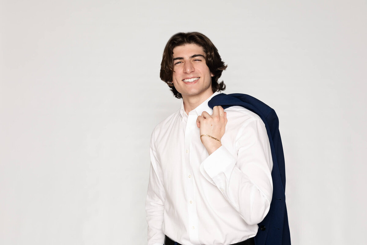 A man with medium-length dark hair is smiling and holding a navy blazer over his shoulder. He is wearing a white dress shirt and standing against a plain light gray background.