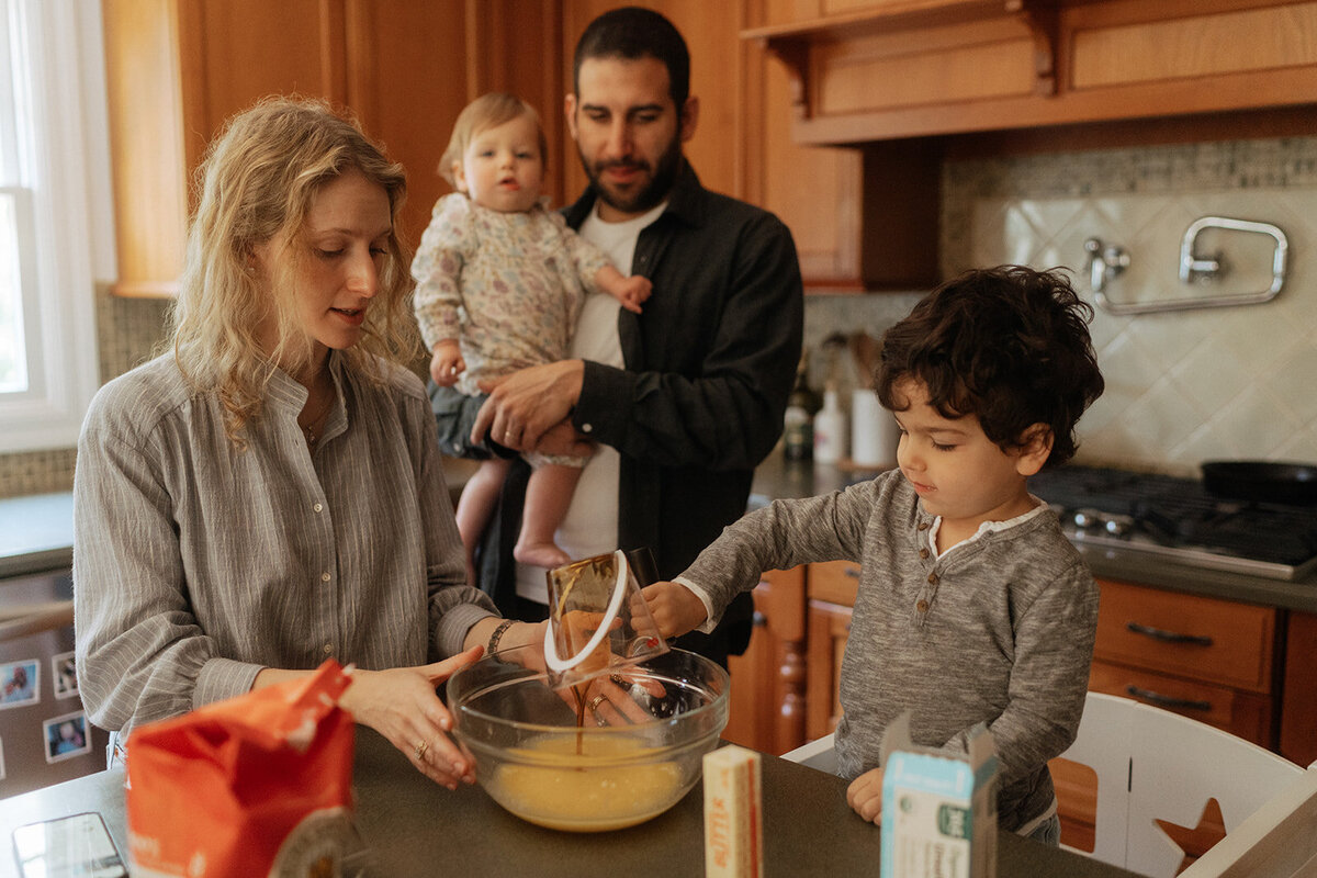 mom makes food with toddler while dad holds baby and watches during family photos captured by NYC family photographer Elsie Goodman 