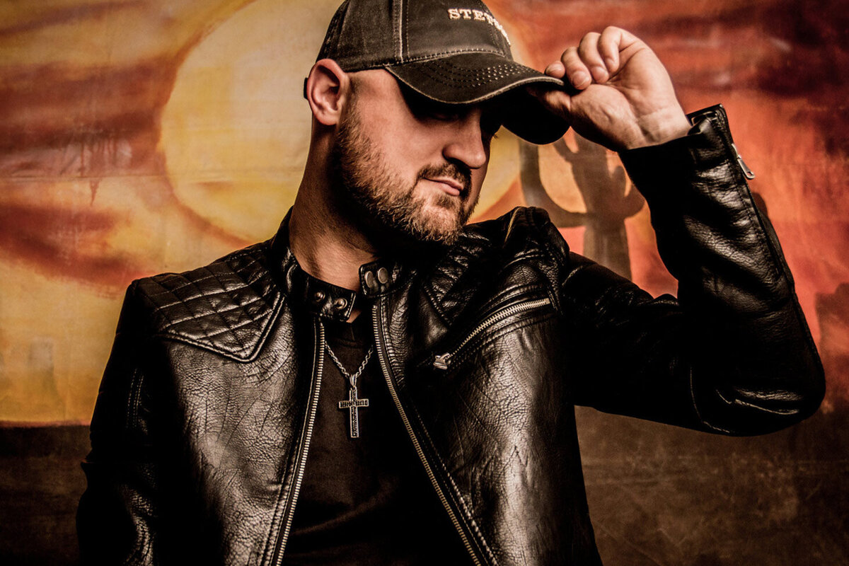Country Music Photo Aaron Goodvin standing in front of wall painted with sun orange sky and cactus wearing black leather jacket and holding brim of baseball cap