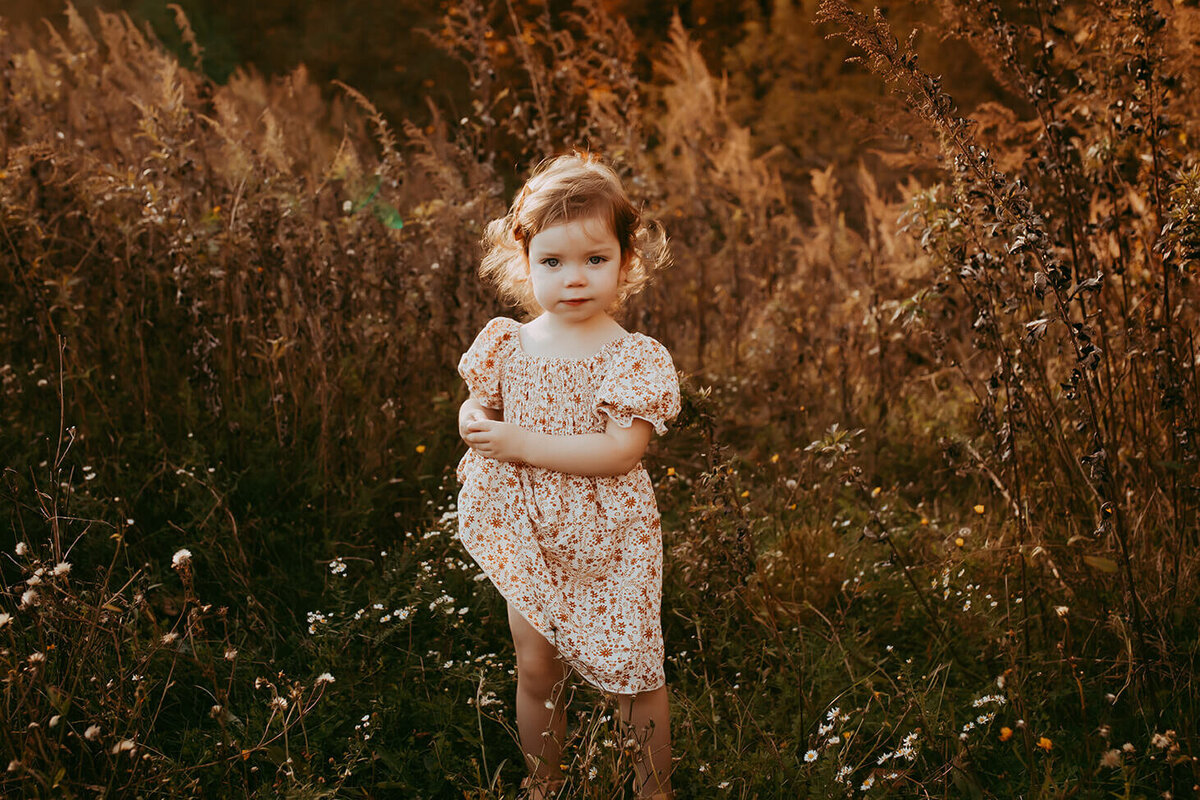 a little girl holding one side of her dress looking serious at the camera in a field