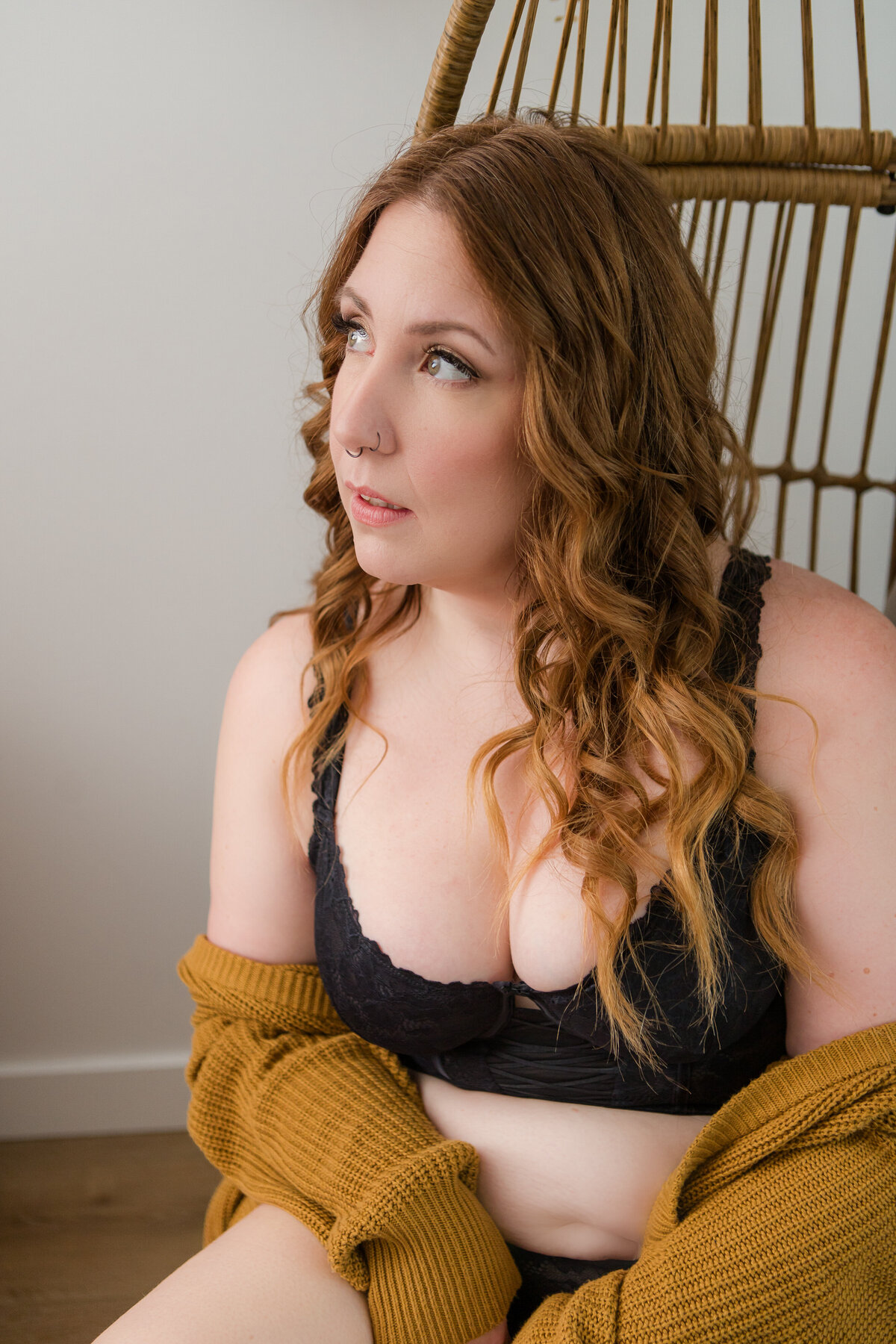 A woman wearing a black bra and a mustard cardigan, sitting in a wicker chair, gazing off in contemplation.