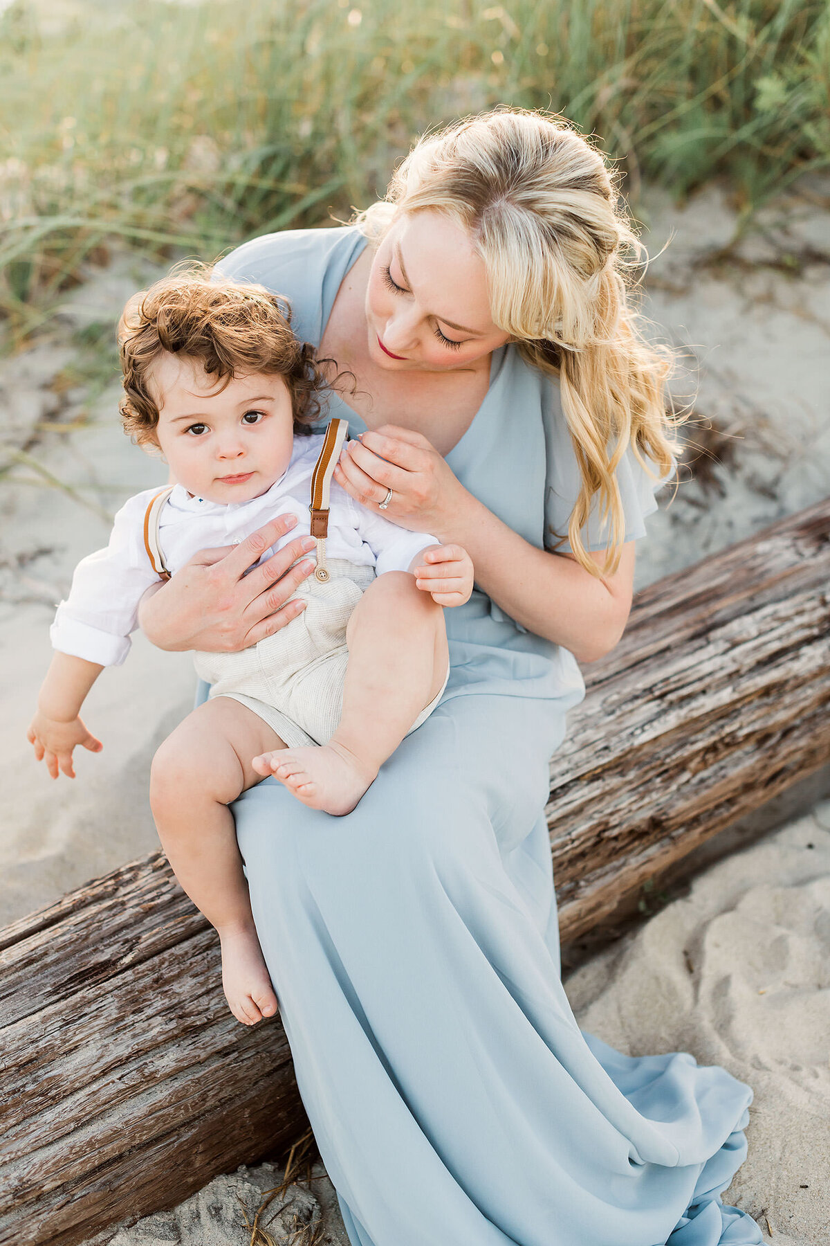 a mom in a blue dress is holding her her son while sitting on a tree stump at the beach