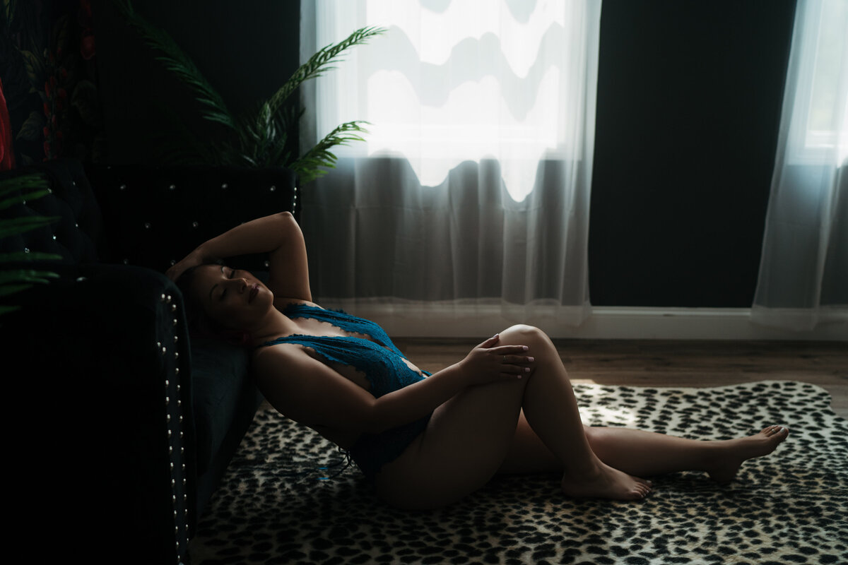 A woman wearing blue lace lingerie sits on a cheetah rug while leaning back onto a black couch in front of windows