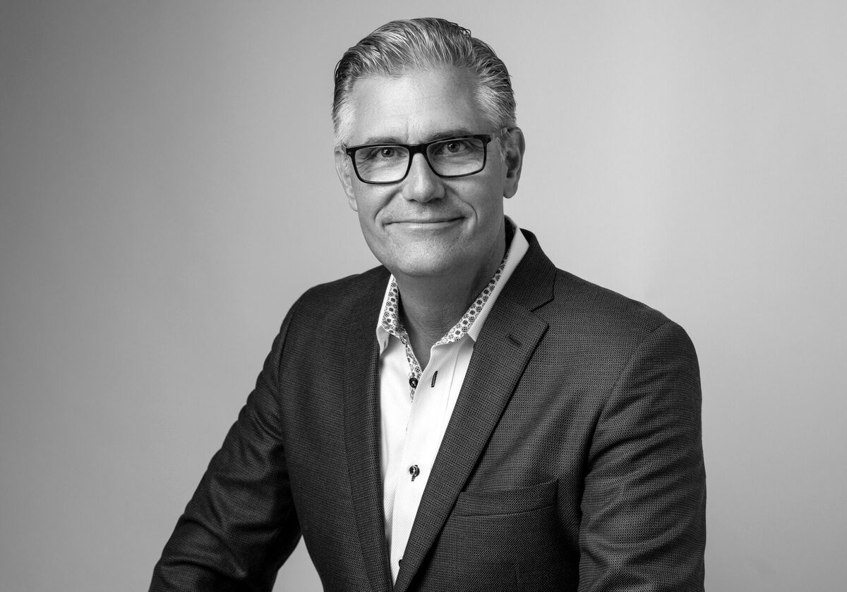 A man with silver hair, wearing glasses and a suit, smiling in a well-lit studio setting.