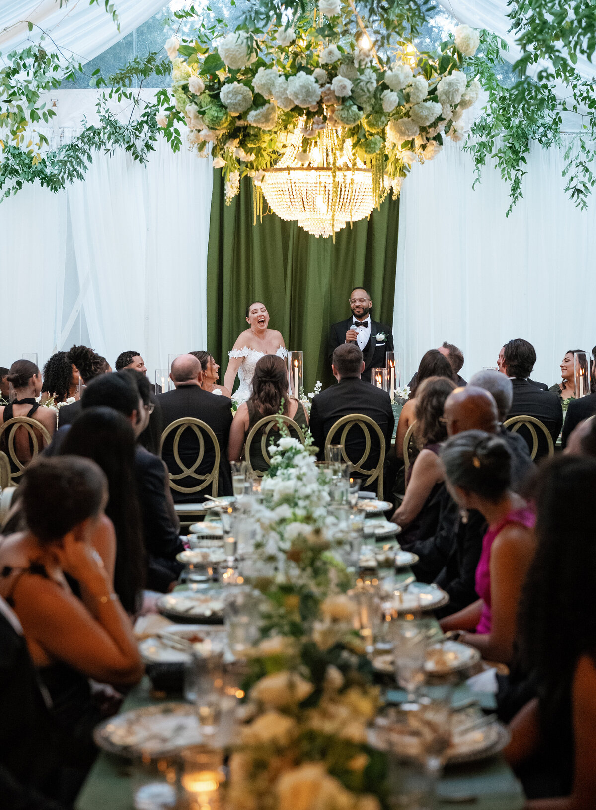 Bride and groom sharing a joyful moment during reception speeches beneath floral chandeliers in a green and white tented reception.