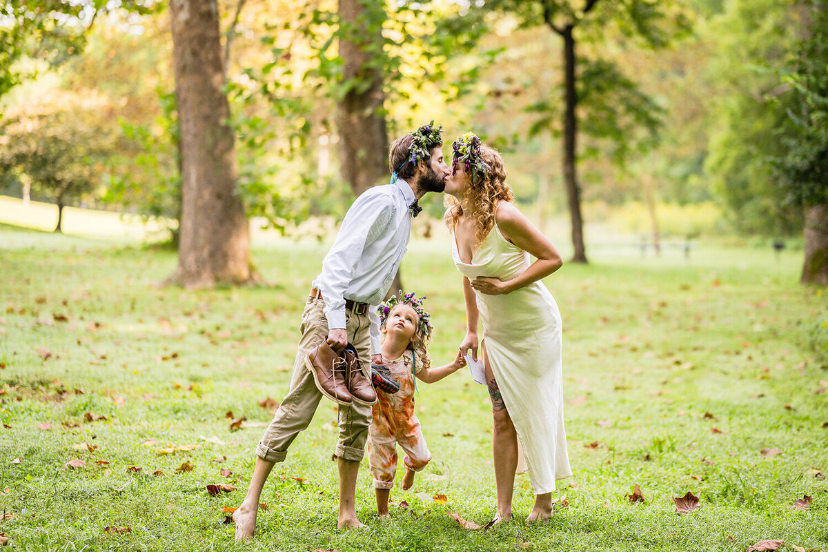 A couple on their elopement day hold hands with their toddler and pause to go in for a kiss while their child observes.