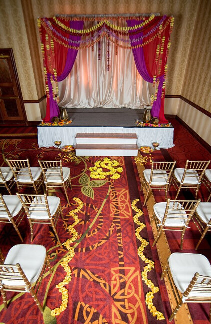 Gold chivari chairs with white cushions line the aisle with yellow rose petals on a red and gold carpet. A red and magenta mandap is decorated with hanging garlands of carnations in Nashville, TN for an Indian Wedding at Embassy Suites