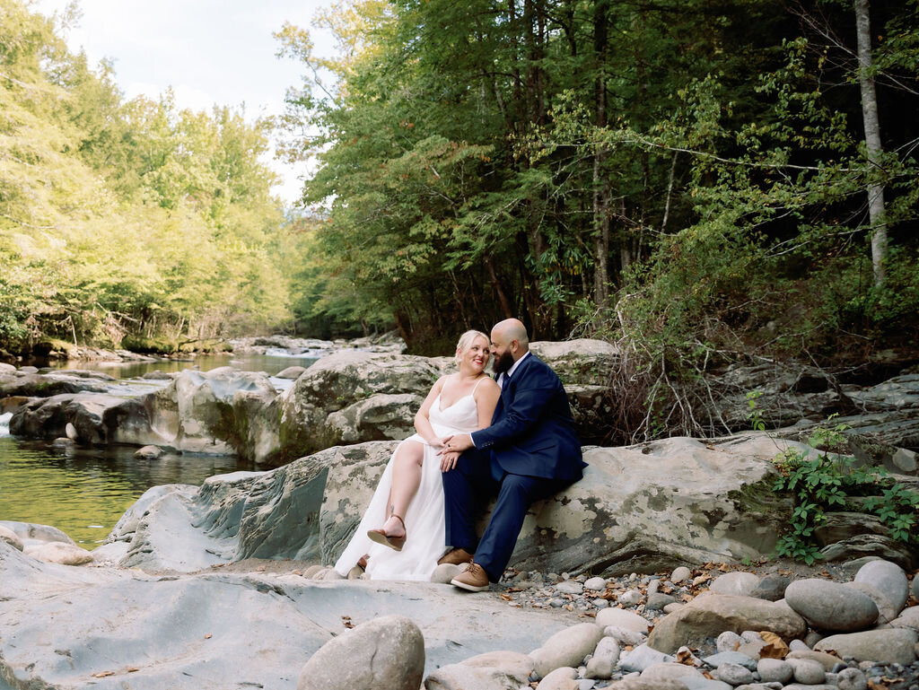 Bride and groom sitting close on a smooth rock by the river at Greenbrier, smiling at each other lovingly during their eloping to Gatlinburg celebration, surrounded by tranquil forest and flowing water.
