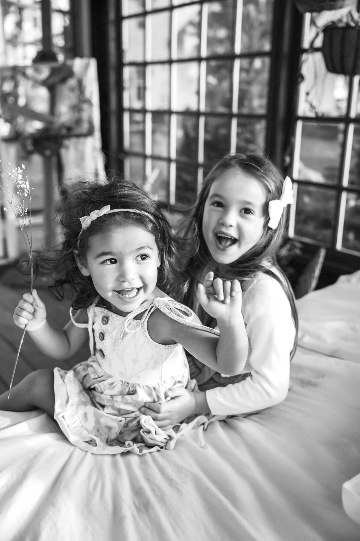 black and white photos of two girls laughing. they are both wearing bows and they are sitting on abed. the younger one is holding a small bunch of tiny flowers