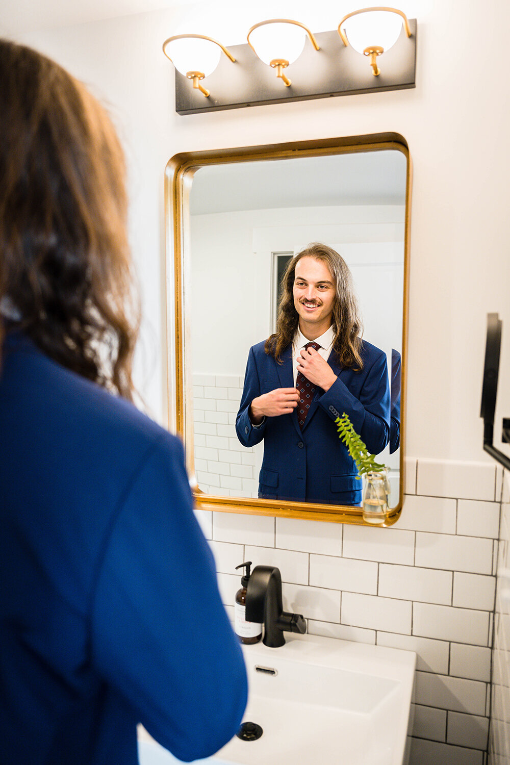 A marrier on his elopement day adjusts his tie in the mirror of a clean and modern hotel bathroom.