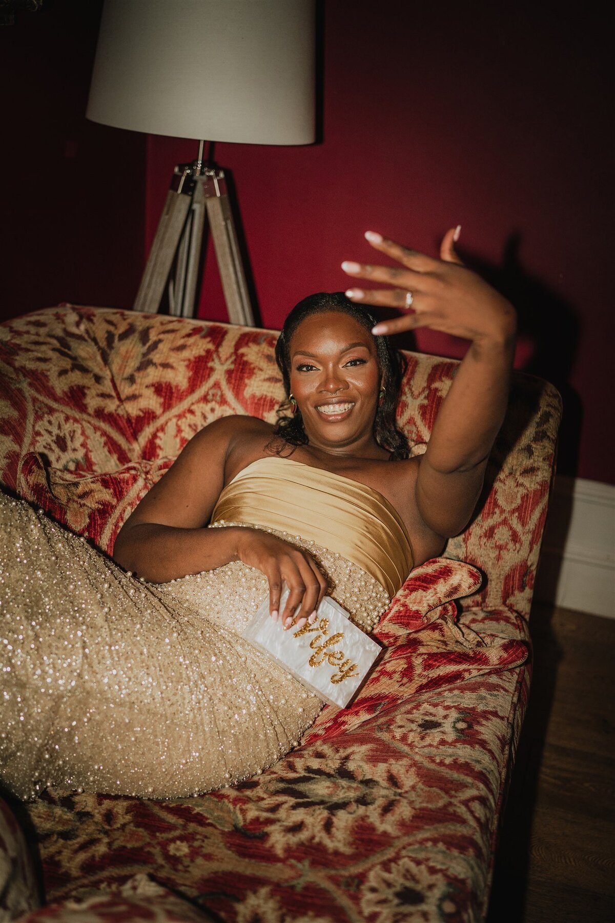 A bride wears a golden sparkly dress and reclines on a sofa. The sofa has a bold pattern and the surrounding walls are red. The bride holds her hand up towards the camera to show off her new wedding ring. She holds a opal textured bad that reads “wifey”.