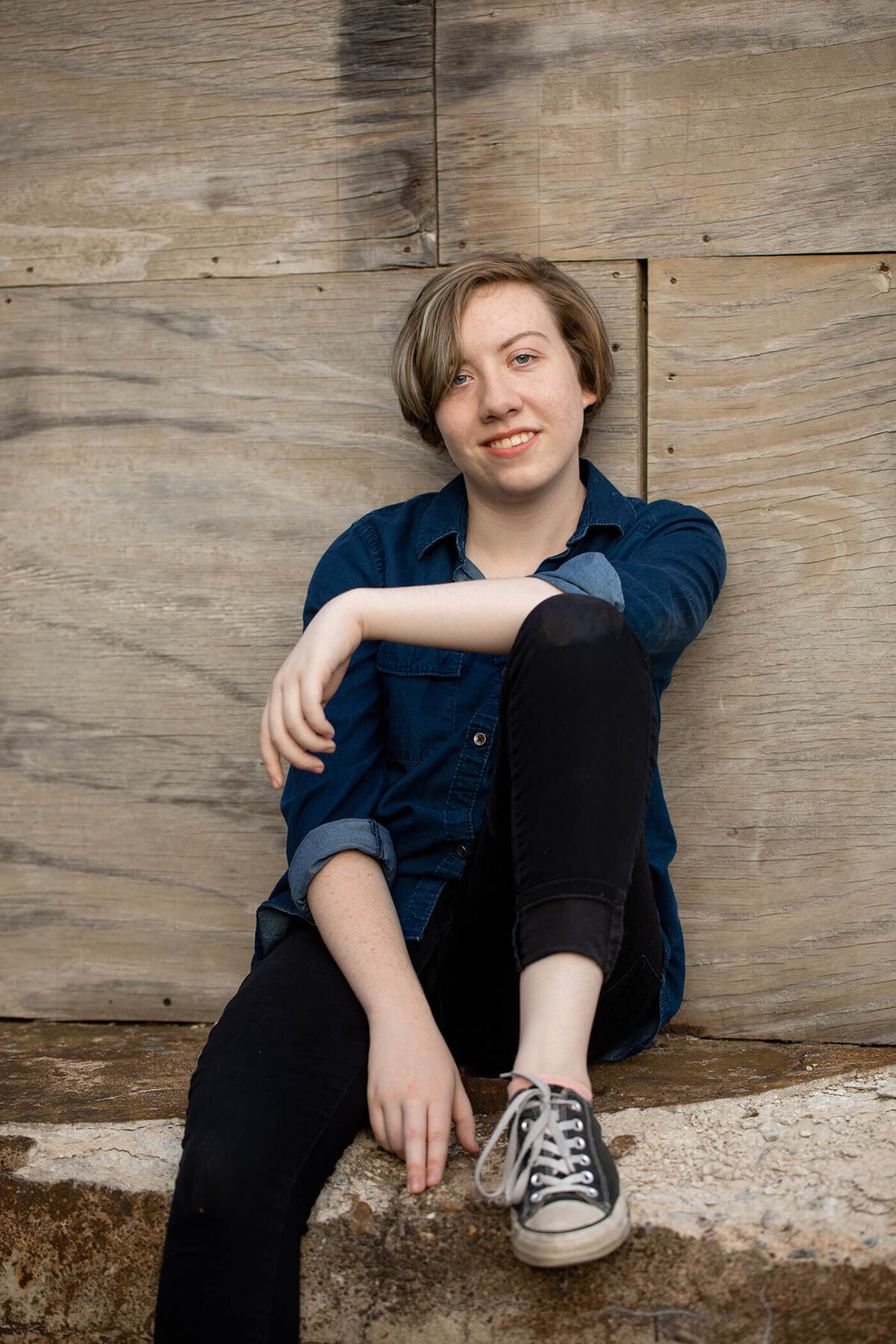 Senior girl wearing a blue top sitting on a ledge with wood background.