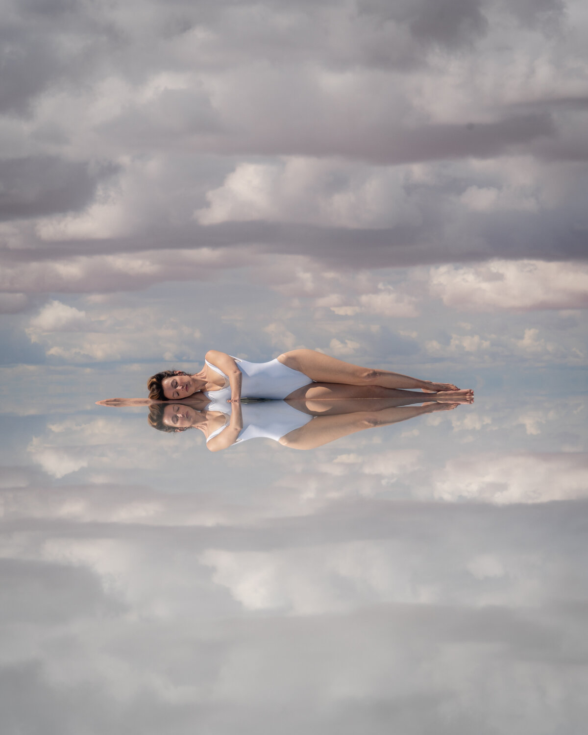 Woman in white bathing suit lying on water with reflection of woman and clouds