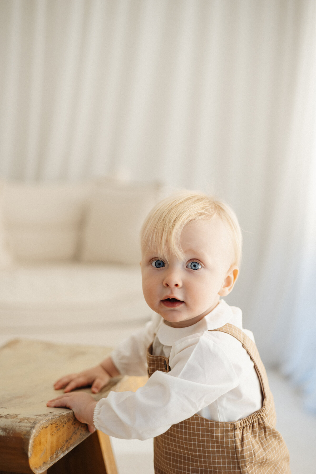 A little boy with blonde hair looking at the camera while holding onto a wooden chair in a neutral styled milestone photoshoot