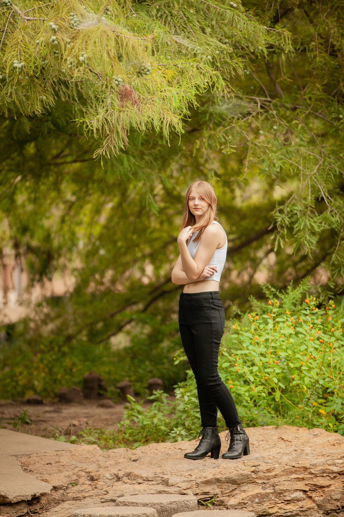 senior girl on rock with green brush and green tree in background playing with hair