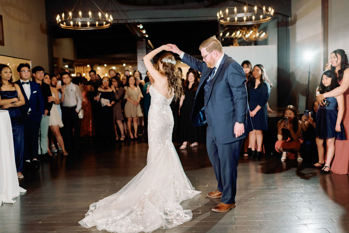 A warm and intimate wedding reception at The Lake House in Calgary, viewed from outside through large glass windows. Guests are seated at long, candlelit tables, engaged in conversation and enjoying the elegant atmosphere. The rustic charm of the venue is highlighted by stone walls, a grand fireplace adorned with greenery and candles, and antler chandeliers casting a soft glow. The dim lighting and reflections in the glass add depth to the scene, creating a cozy and romantic ambiance for the celebration.