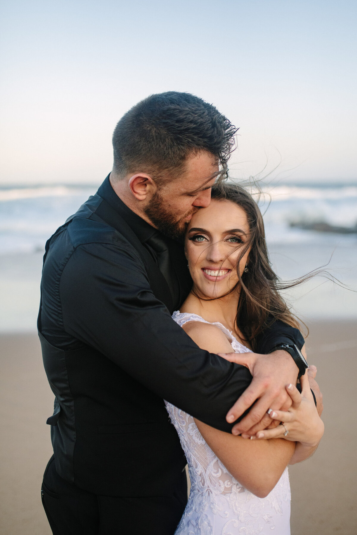 A groom wraps his arms around his bride while giving her a soft kiss on her forehead. 