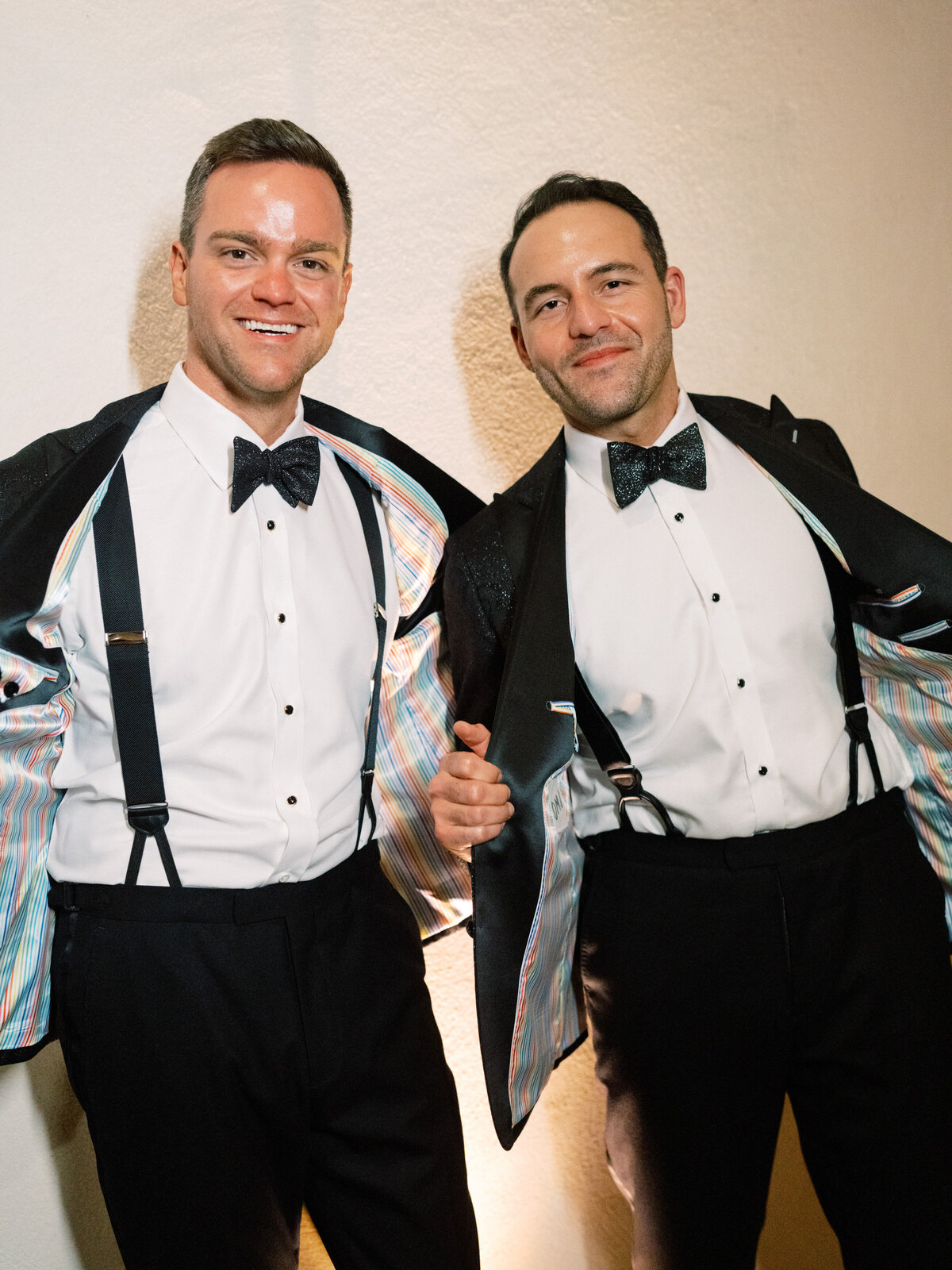Two well-dressed men in tuxedos proudly display their colorful jacket linings, capturing a moment of joy and style at a formal event surrounded by elegance and celebration.