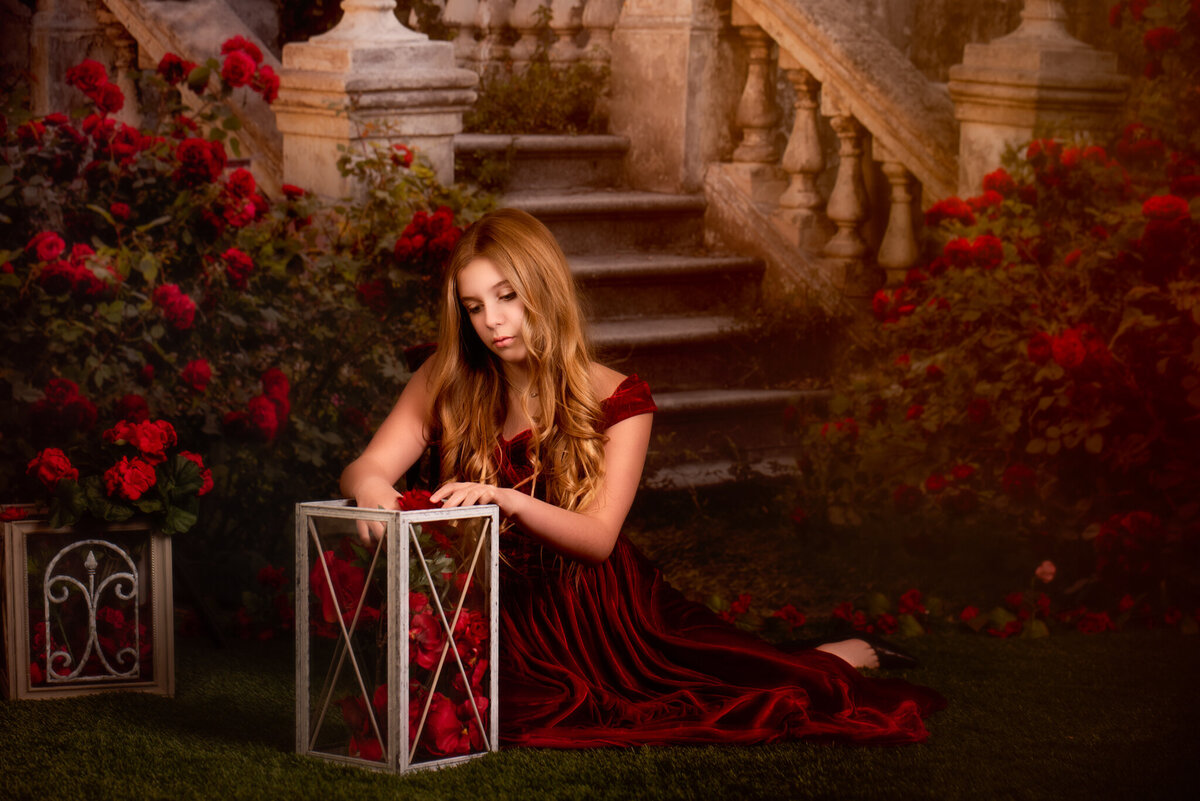 Whimsical fine art portrait of a young girl in a deep red gown, holding a glowing lantern surrounded by vibrant roses. Captured in Overland Park, Kansas, this enchanting setup is ideal for families seeking creative and magical child photography