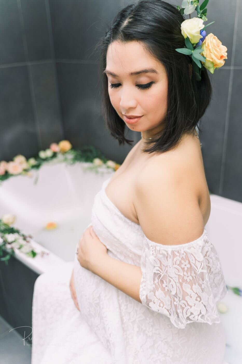 Pregnant woman with floral hairpiece sitting by the milk bath and softly gazing down at her baby bump while wearing an off-shoulder lace dress.