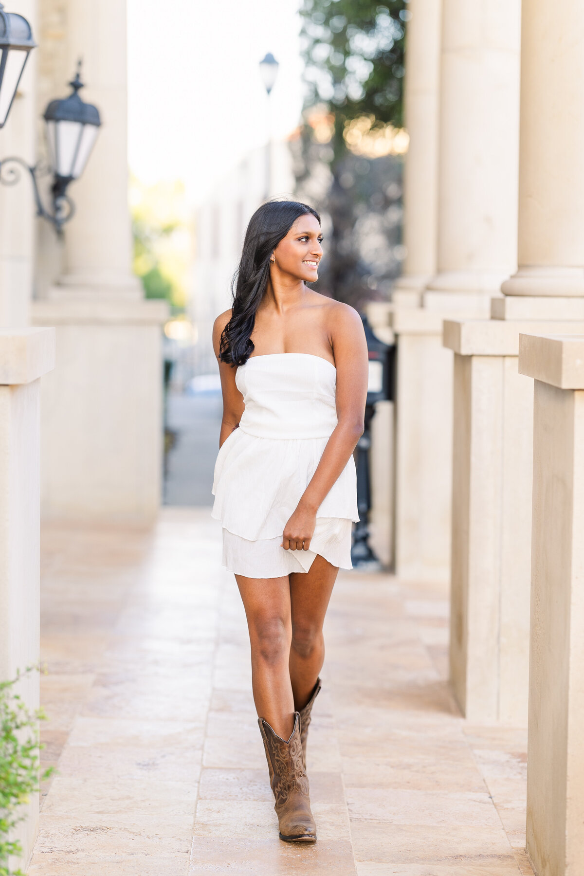Senior girl walking confidently in a white strapless dress and cowboy boots at Adriatica Village. Surrounded by elegant stone columns and soft morning light, she smiles while glancing to the side.