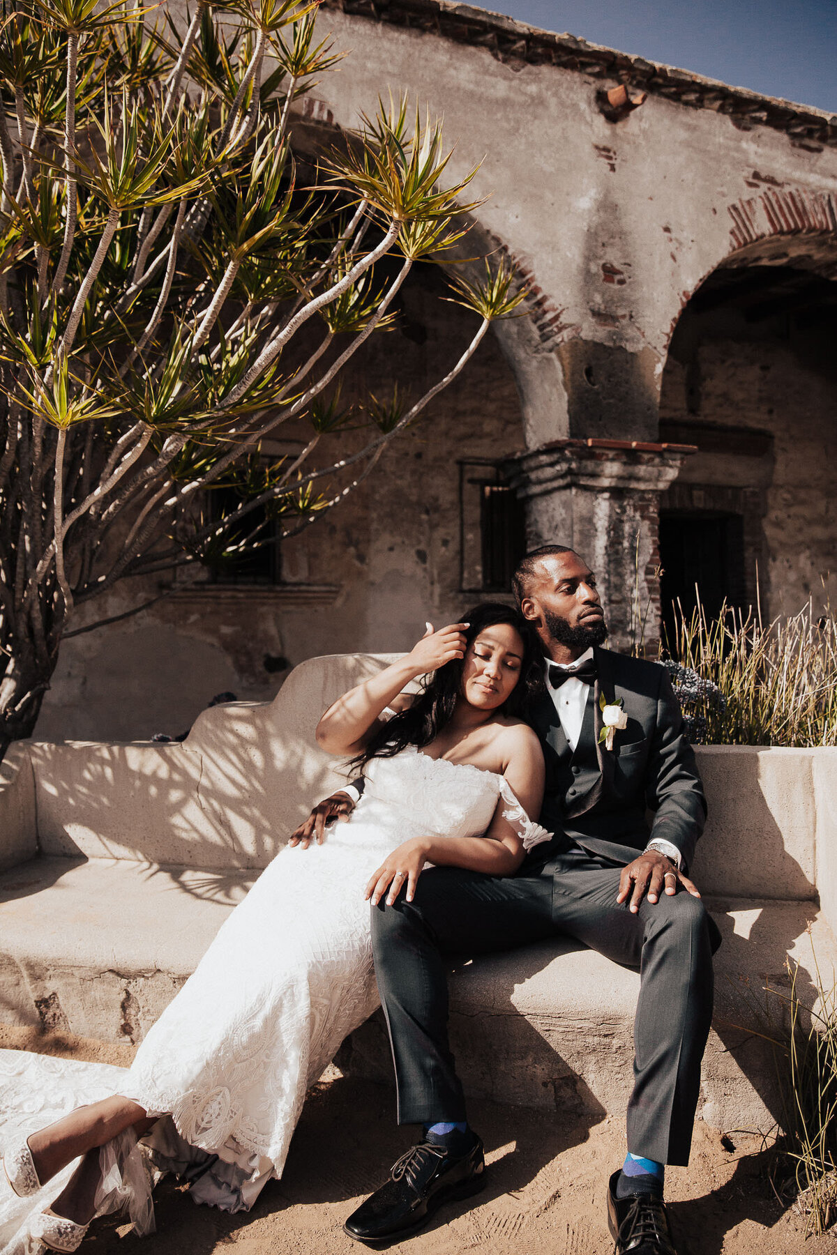 Bride and Groom closely sitting on stone bench in front of Wife portraits // Mission San Juan Capistrano ruins in California