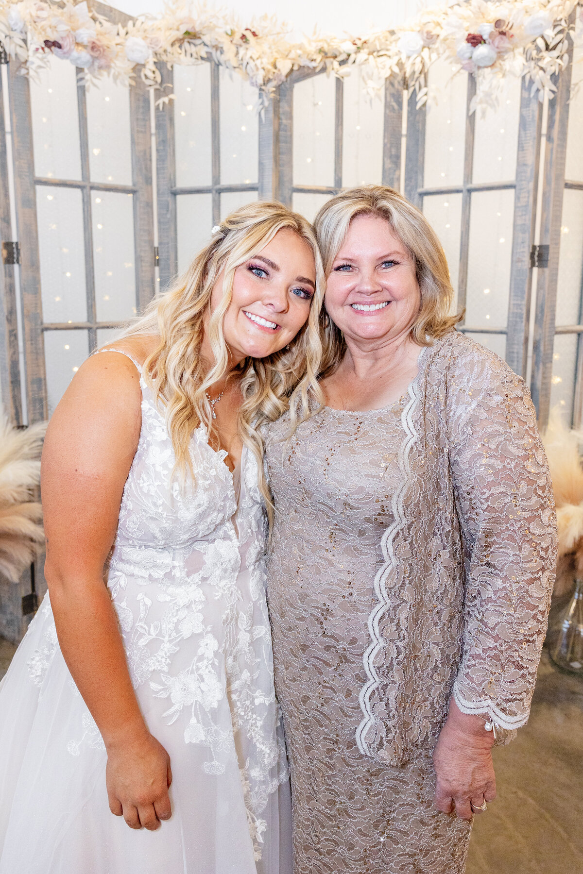 The bride and her mother share a close, heartfelt moment, smiling at the camera in front of the rustic wedding backdrop. Both women radiate joy, love, and elegance in their beautifully detailed gowns.