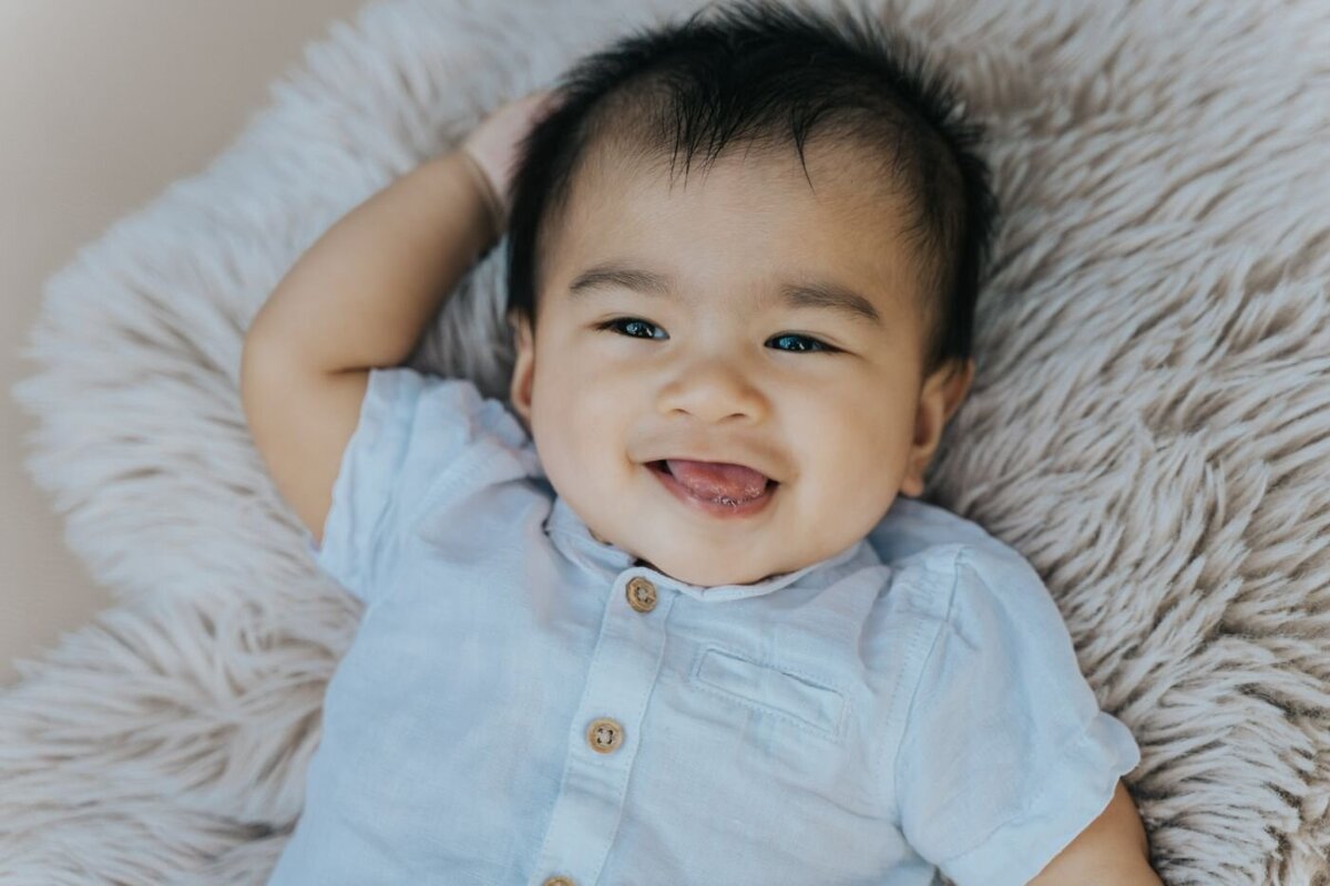Baby boy with dark hair wearing a light blue shirt, laughing while lying on a fluffy beige blanket.