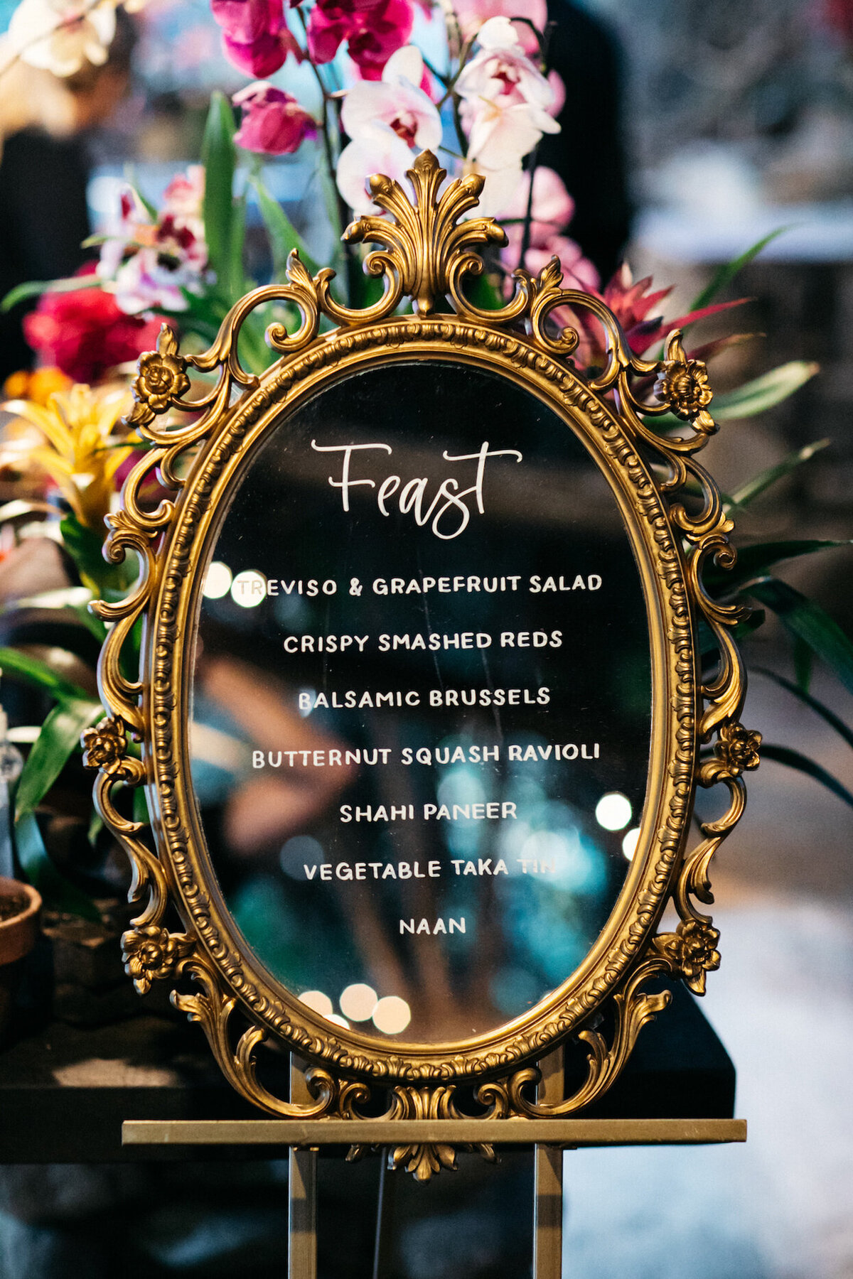 Ornate oval mirror with menu for a wedding reception, with colorful orchids in the background
