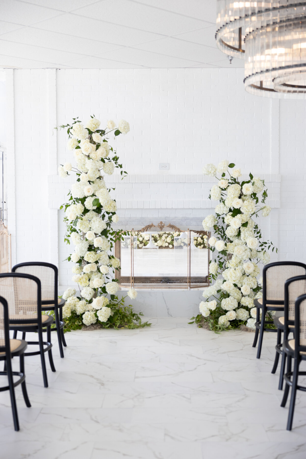 Elegant indoor wedding ceremony setup with black rattan chairs facing a white floral arch arrangement in front of a fireplace.