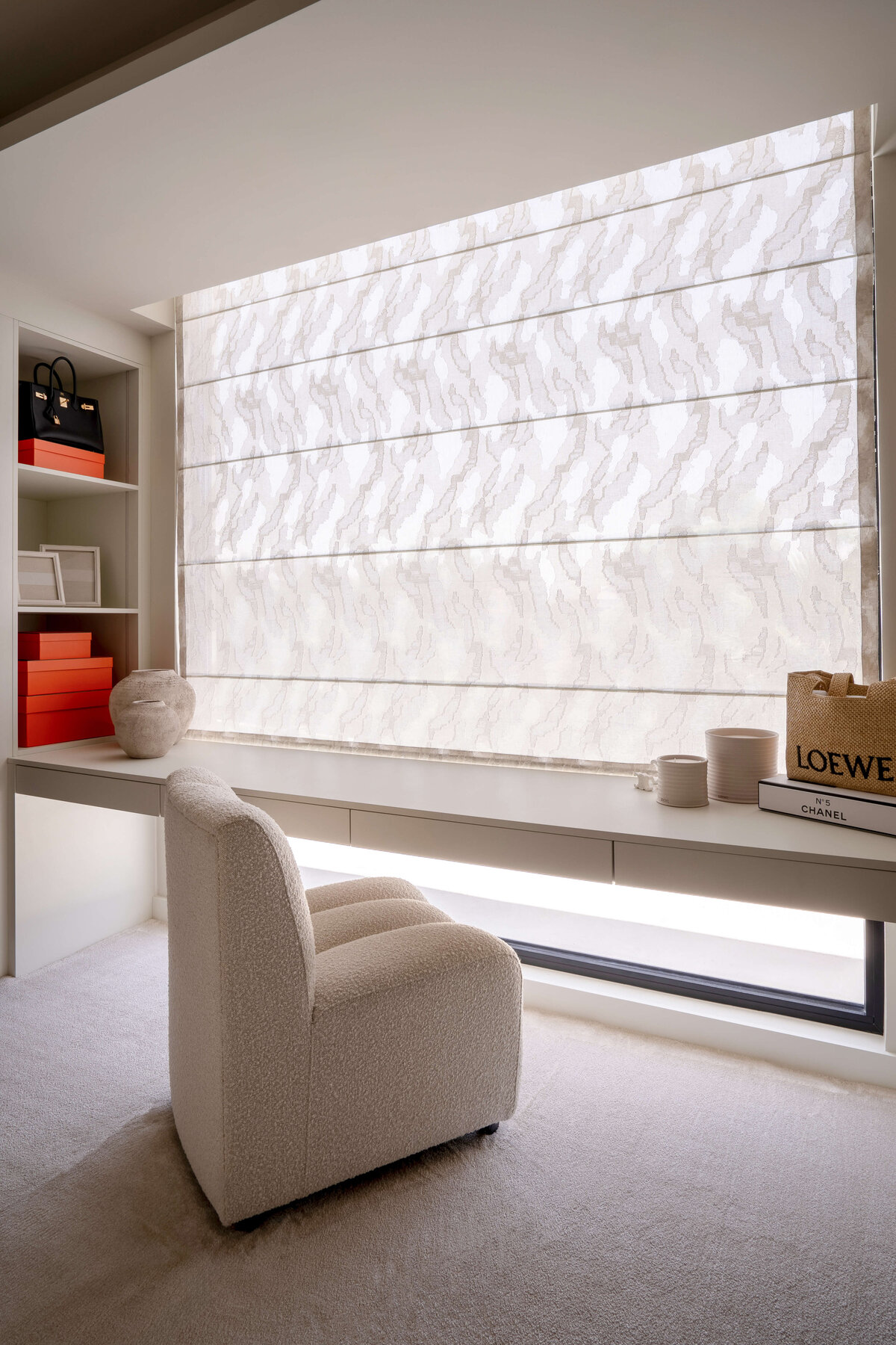 Desk on the far wall of the primary dressing room. The desk is white with a soft, textured white chair pulled up. There are built-in shelves to the left with orange boxes.