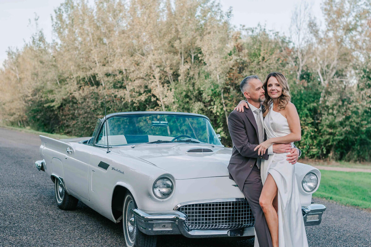 Bride and groom sit on the hood of a car. Bride looks at me and groom looks at her.