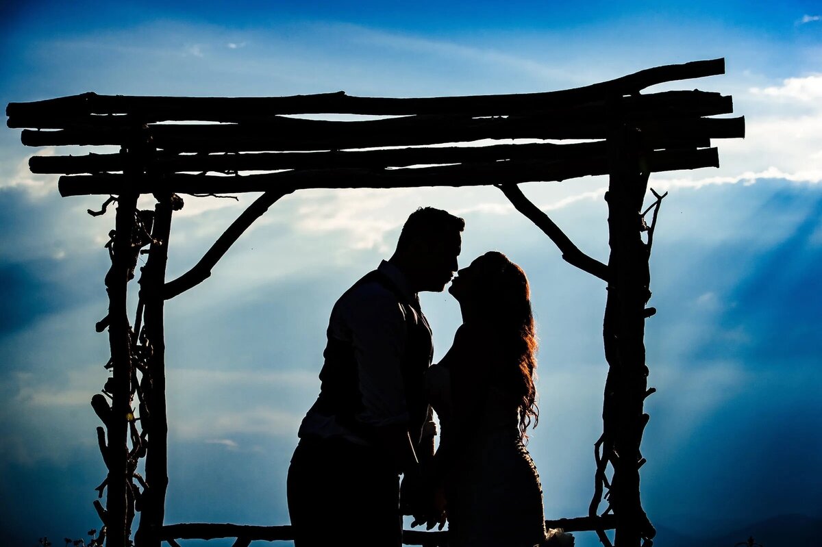 A silhouette of a couple sharing an intimate moment under a rustic wooden arch, with the sun's rays creating a dramatic backdrop.