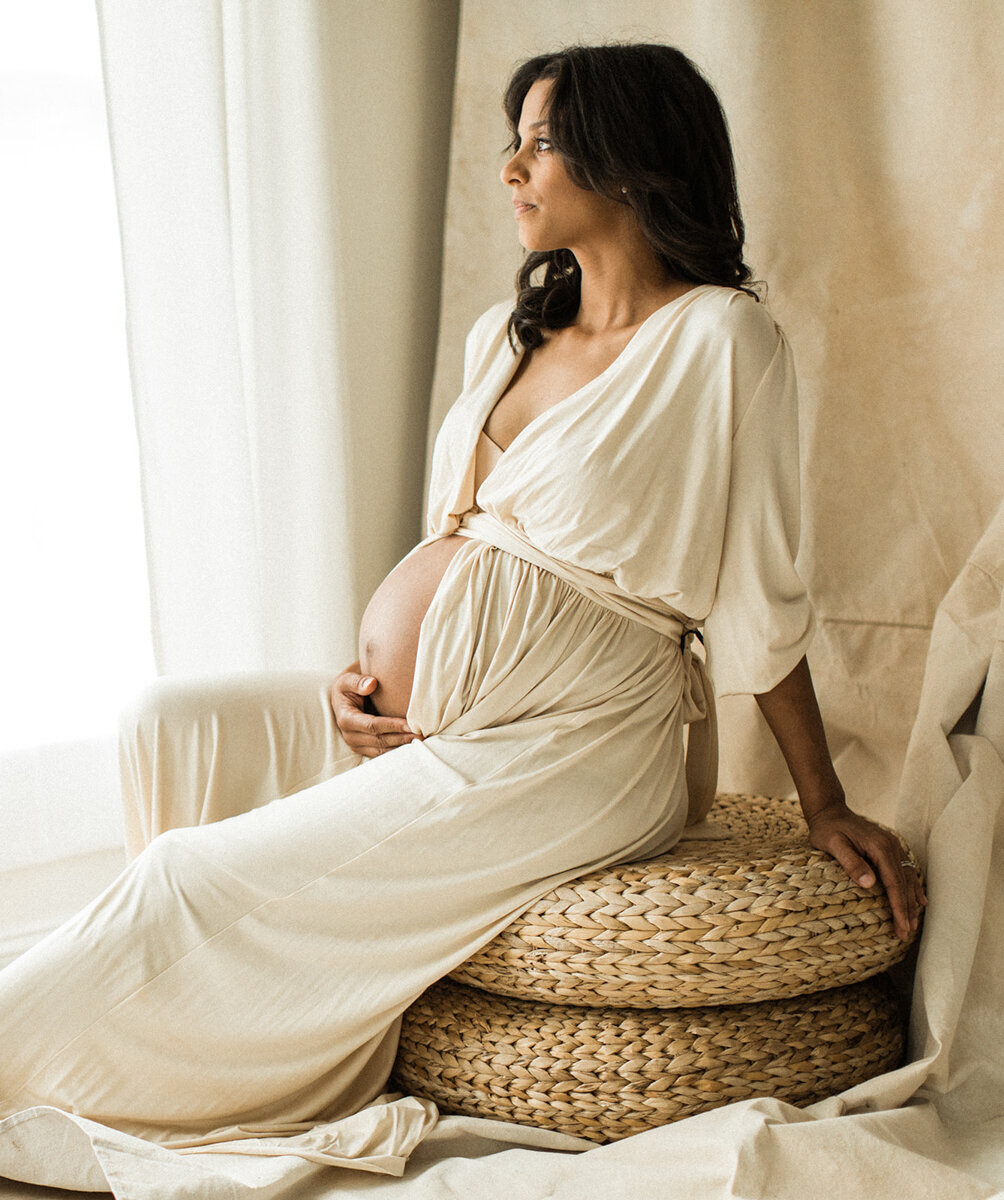 studio maternity portrait of woman of color sitting on wicker seat with cream backdrop by window