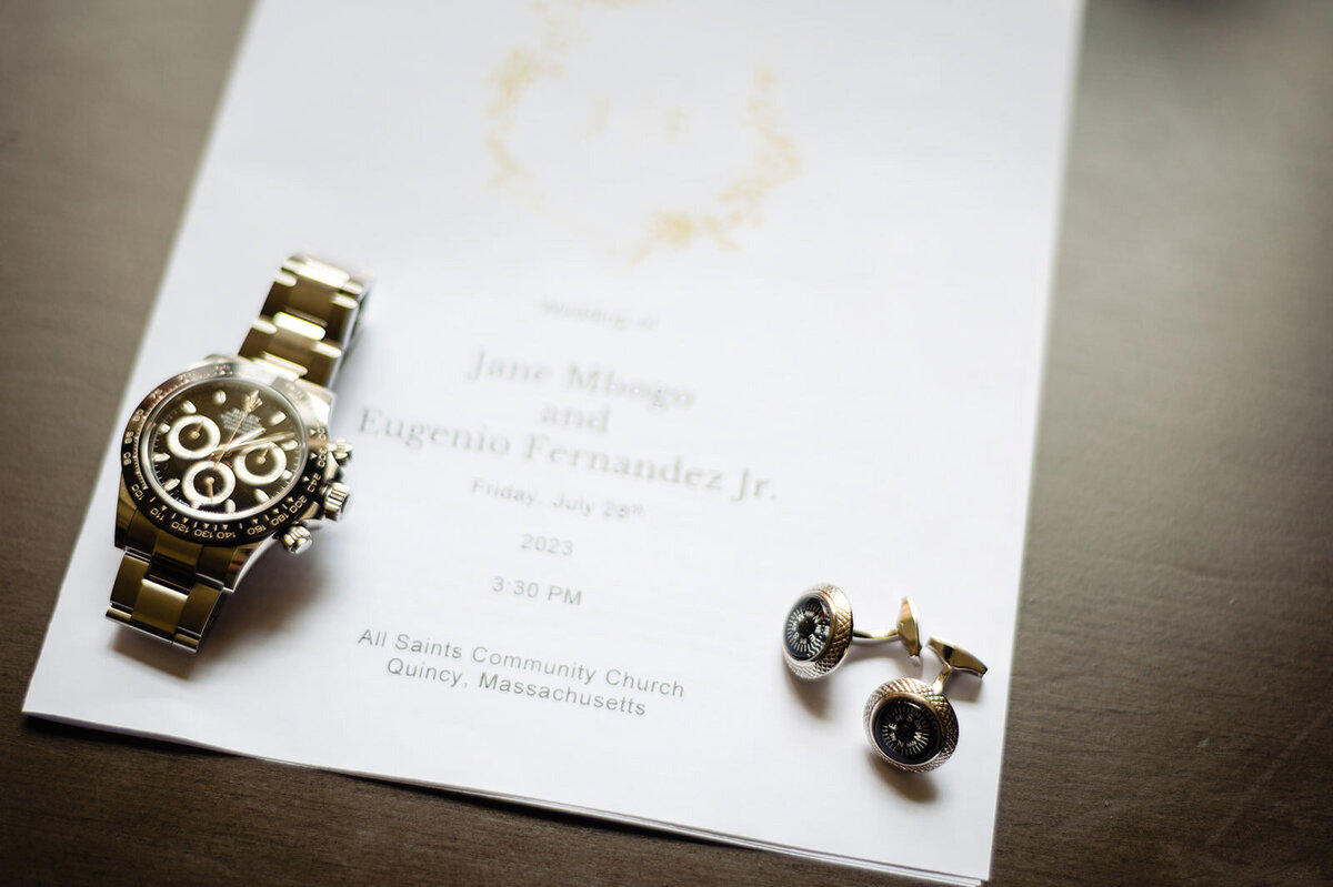 A wedding invitation lying next to a luxury wristwatch and a pair of cufflinks on a textured surface, detailing the names 'Jane Masego' and 'Eugenio Fernandez Jr.' with the date and location of the wedding in Quincy, Massachusetts.
