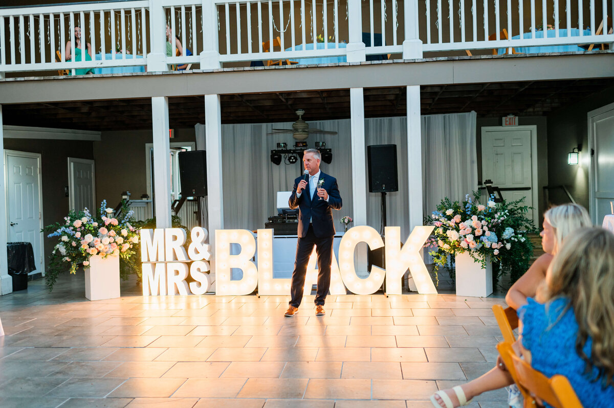 A man in a suit speaks into a microphone at an outdoor event. Behind him, large illuminated letters spell out “MR and MRS BLACK.” Flowers decorate the area, and guests are seated at tables in the foreground.
