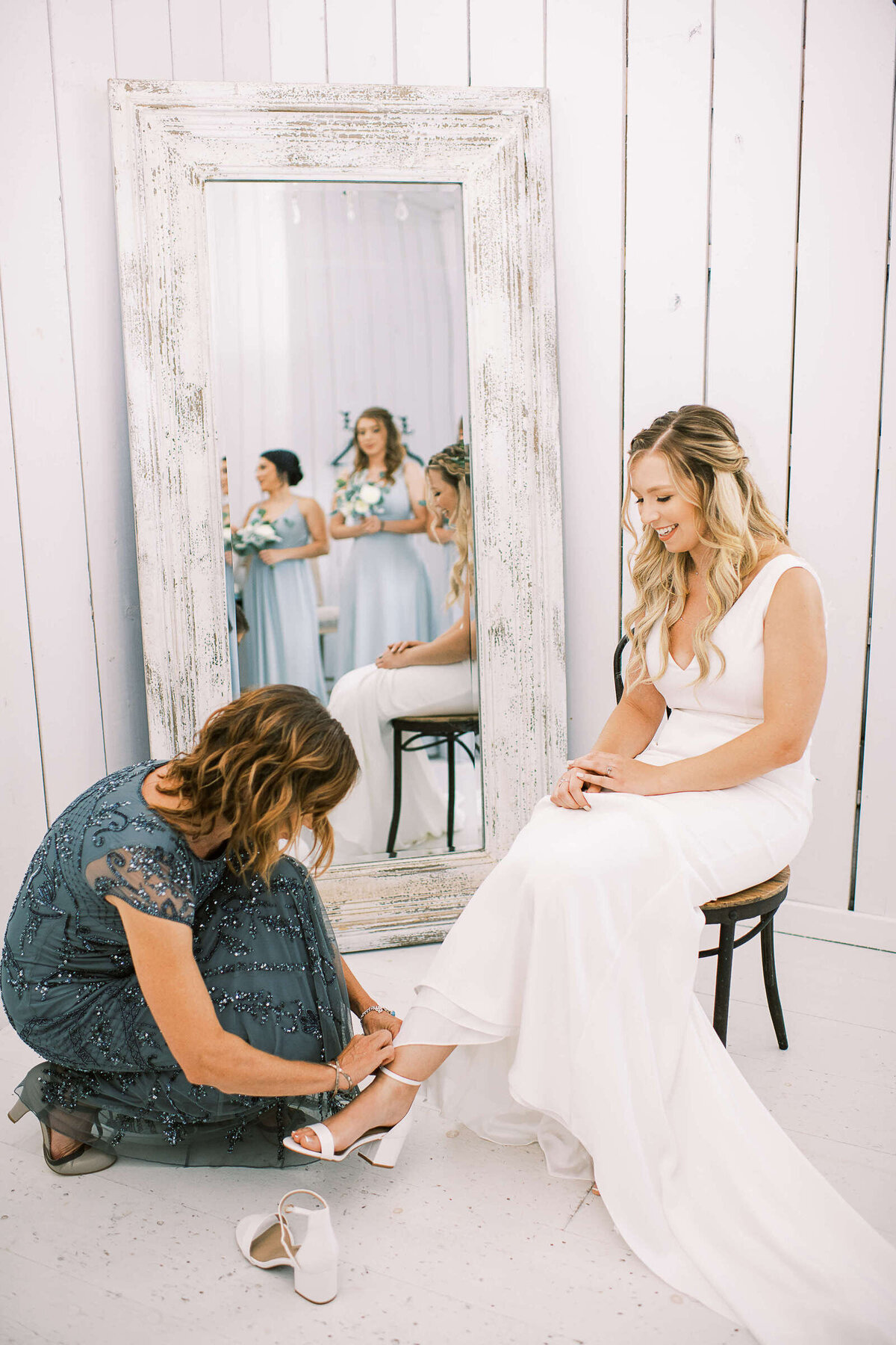 Mother of the bride helps bride put on her white wedding heels