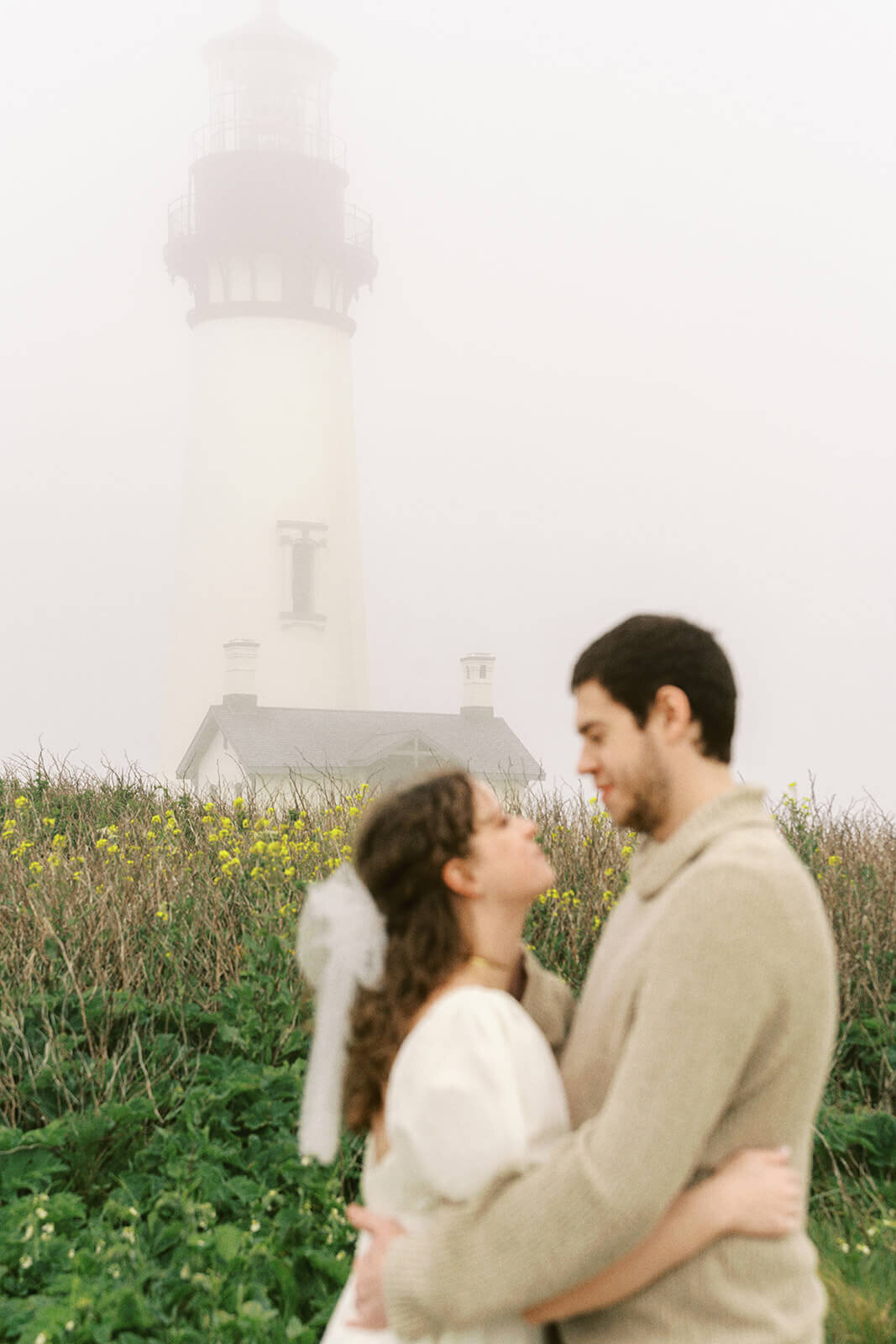 A couple are out of focus while embracing with Yaquina Head Lighthouse in focus behind them. 