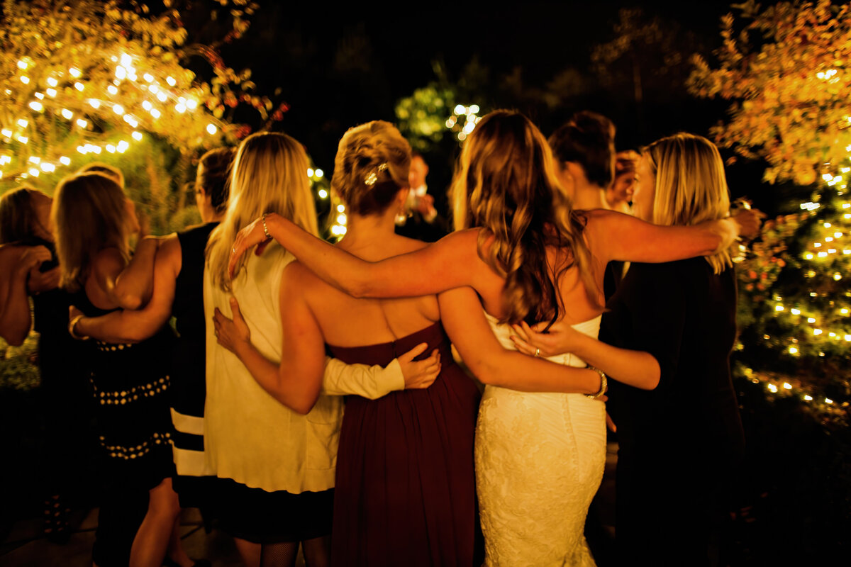The bride and her friends are seen from behind with their arms around each other at night.