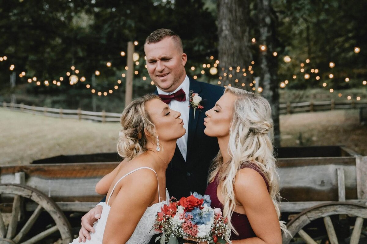 A groom stands behind two women about to kiss, with string lights and a vintage wagon in the background.