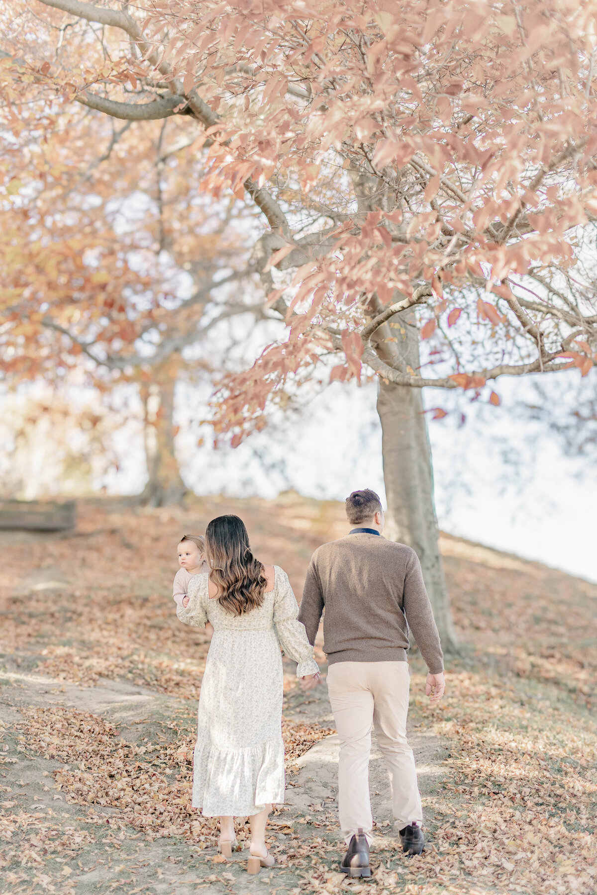Parents holding hands and walking up a hill covered in fallen leaves during their daughter’s first birthday session in a Kansas City park.