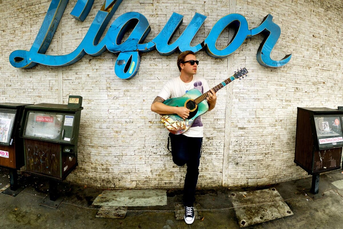 Musician photo Santa Monica Daniel Wesley standing in front of liquor store wall news paper boxes on either side of him holding sea foam blue electric guitar Just Play Something Gallery