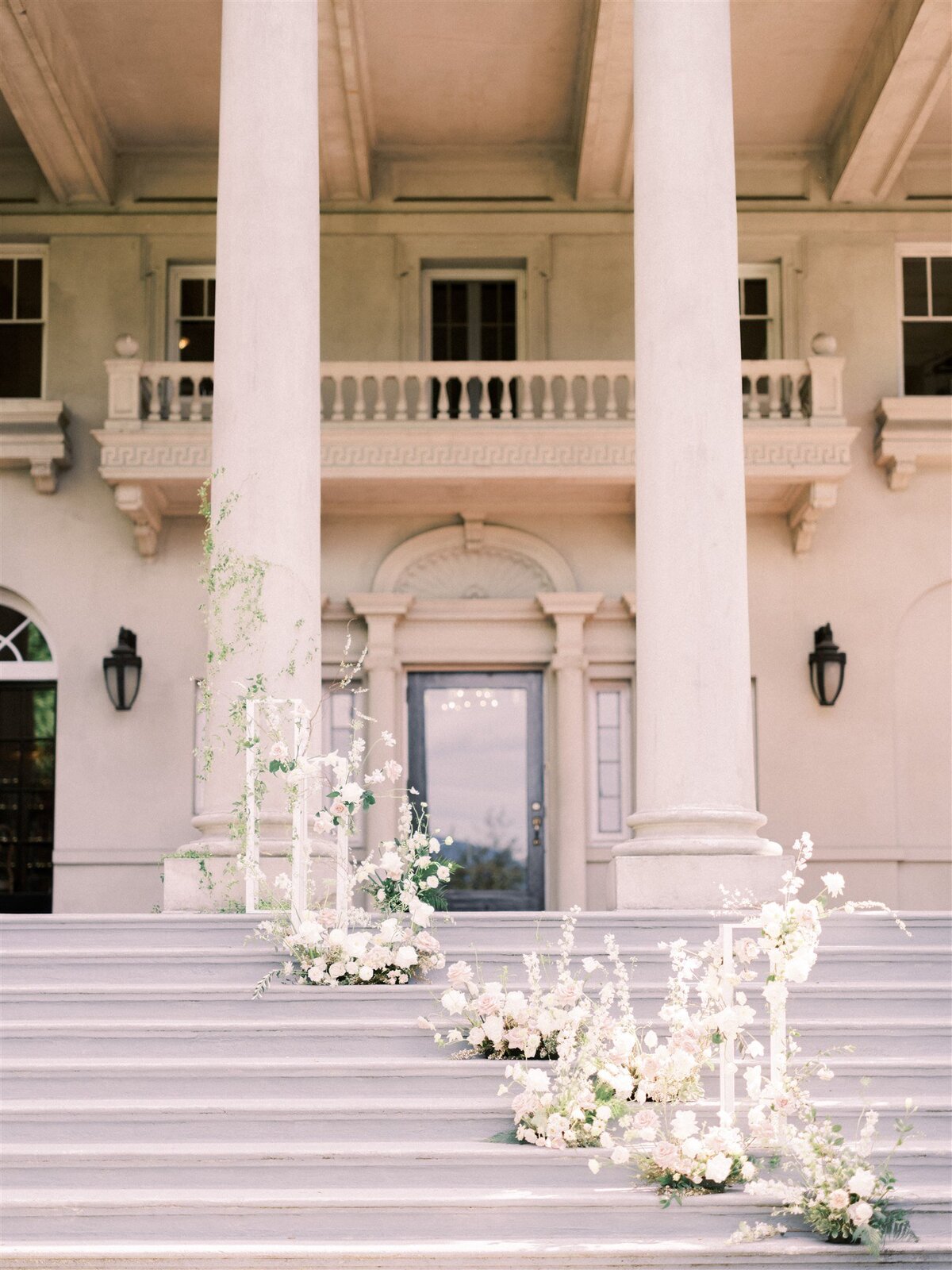 A grand entrance with high pillars, a set of steps adorned with white floral arrangements, leading to a large wooden door on a classical building façade, all meticulously designed by Destination Wedding Planner Melissa Dawn Event Designs.