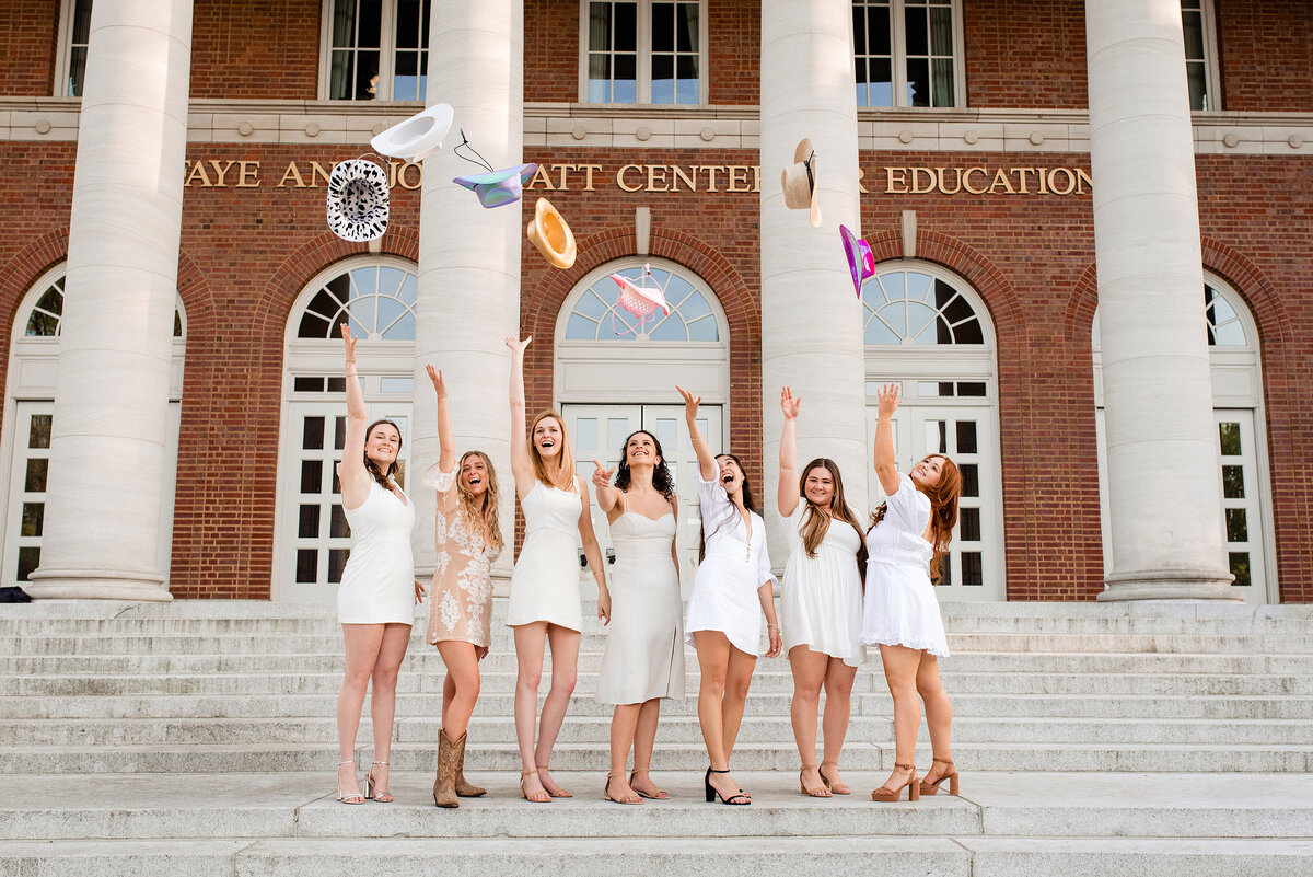 Group of senior girls standing on stairs at Vanderbilt all wearing white dresses and tossing cowgirl hats into the air