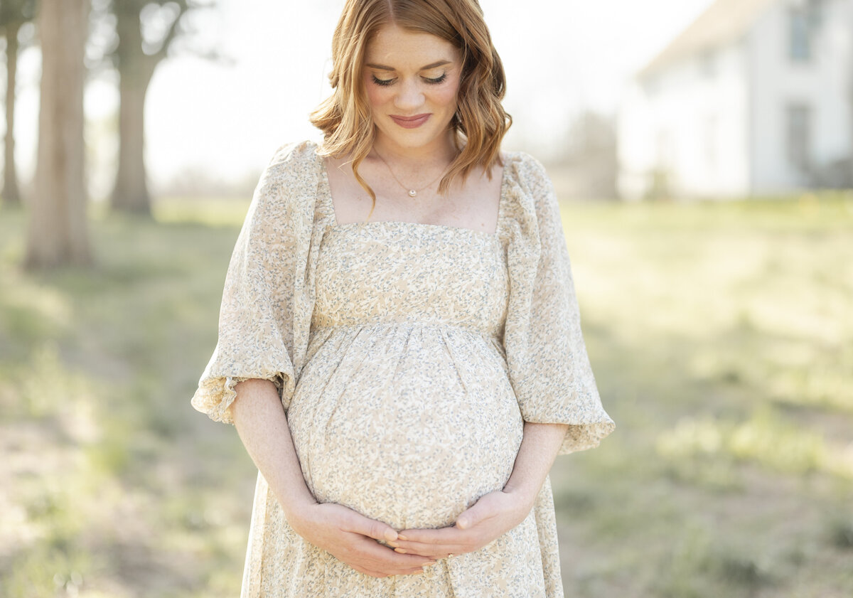 Red-haired pregnant woman in a cream gown holding her bump outdoors during golden hour.