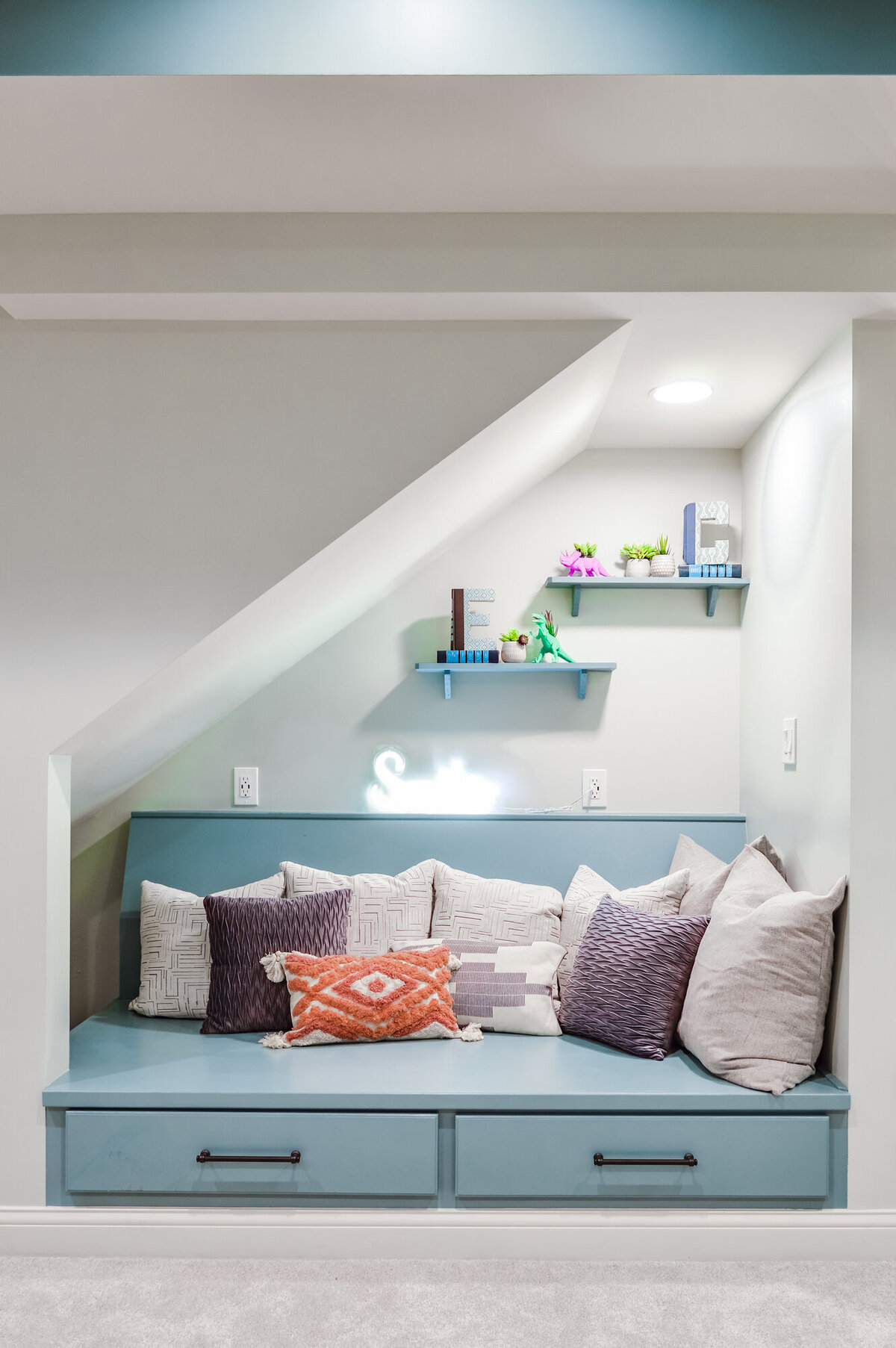 A cozy reading nook built into a corner with light gray walls. A long, light blue cushioned bench with two drawers underneath is lined with numerous throw pillows in various textures and patterns, including stripes, geometric designs, and a colorful southwestern-inspired pillow. Above the bench, two light blue shelves are mounted on the wall, displaying small decorative objects, books, and potted artificial plants. The nook is illuminated by soft lighting, creating an inviting and comfortable space. A light gray carpet covers the floor.