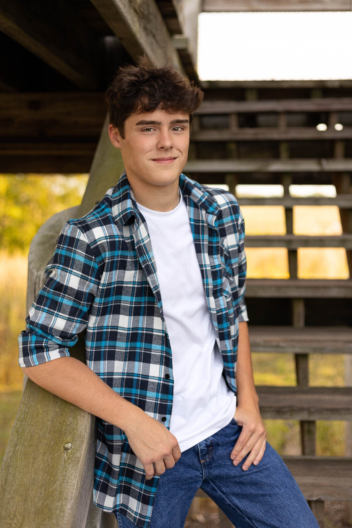 A senior guy standing on wooden stairs leaning on the railing looking at the camera in Lawrence, KS